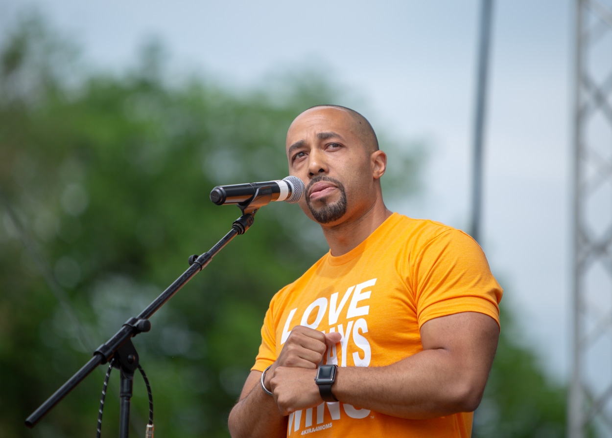 Charles Johnson speaking passionately at outdoor rally in orange Love Always 4Kira4Moms shirt